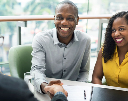 A smiling man shaking hands across a table with a woman in yellow also smiling.