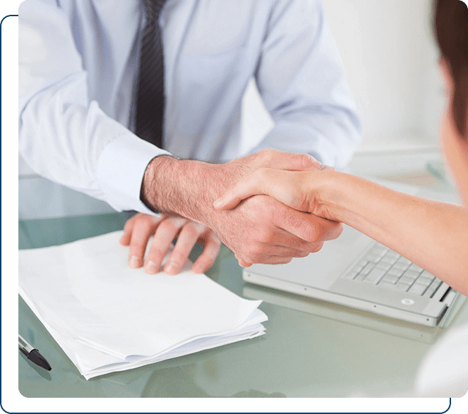 Two professionals shaking hands across a desk, with paperwork and a laptop.